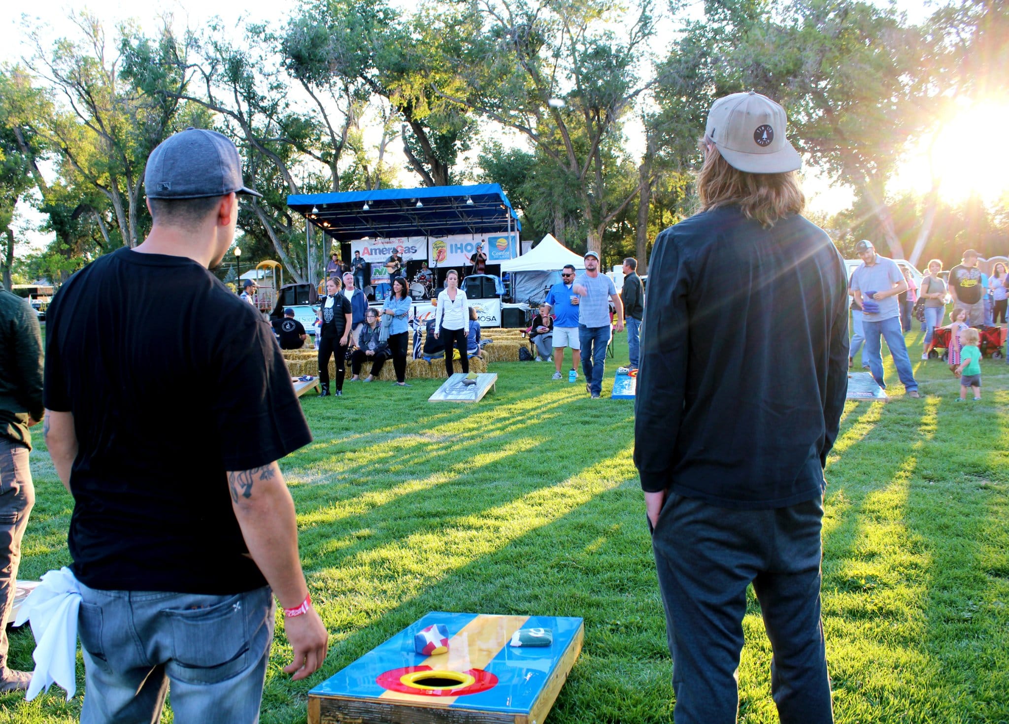 People playing in a cornhole tournament at Beat the Heat BBQ & Brews festival in Alamosa. The sun shining through the trees and casting shadows on the grass inbetween the cornhole boards