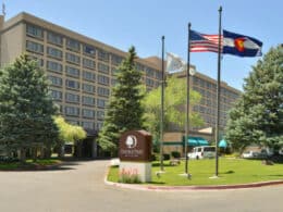 Outside of DoubleTree Grand Junction, a beige multi-story hotel building with 3 flags raised outside- a Colorado state flag, the American flag, and a DoubleTree flag
