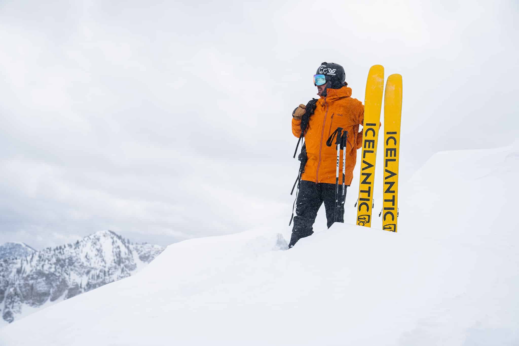 Skier in bright orange jacket standing on top of a snowy mountain next to yellow Icelantic skis