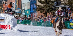 Man on horse pulls a man on skis over a jump built out of snow on the main street of a town
