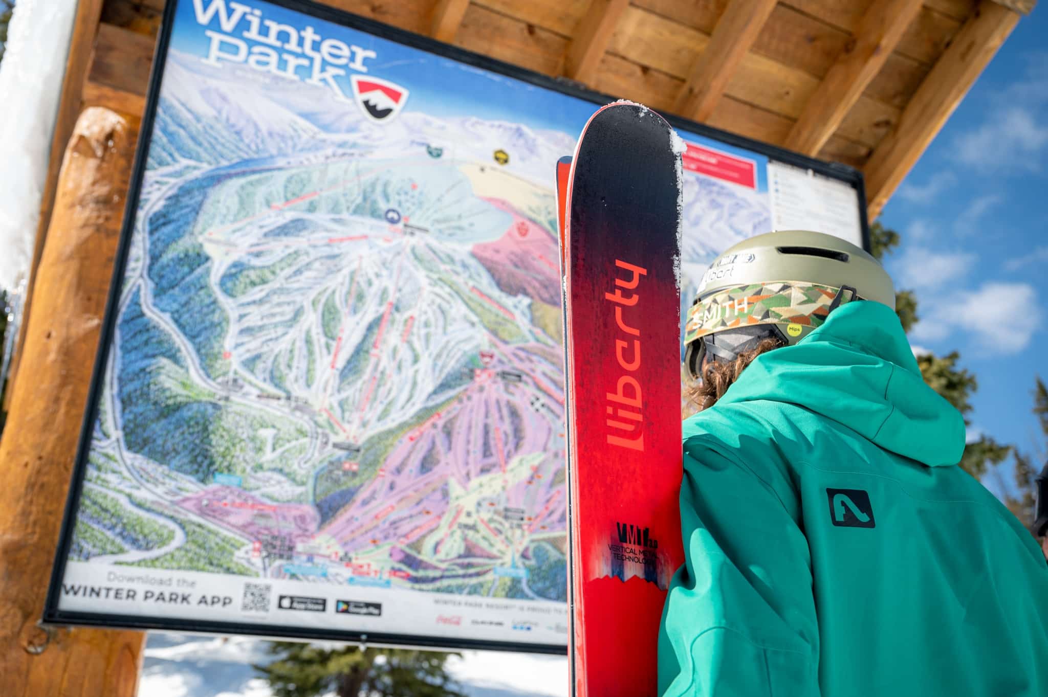 skier in a teal jacket standing in front of Winter Park slope map holding red Liberty skis