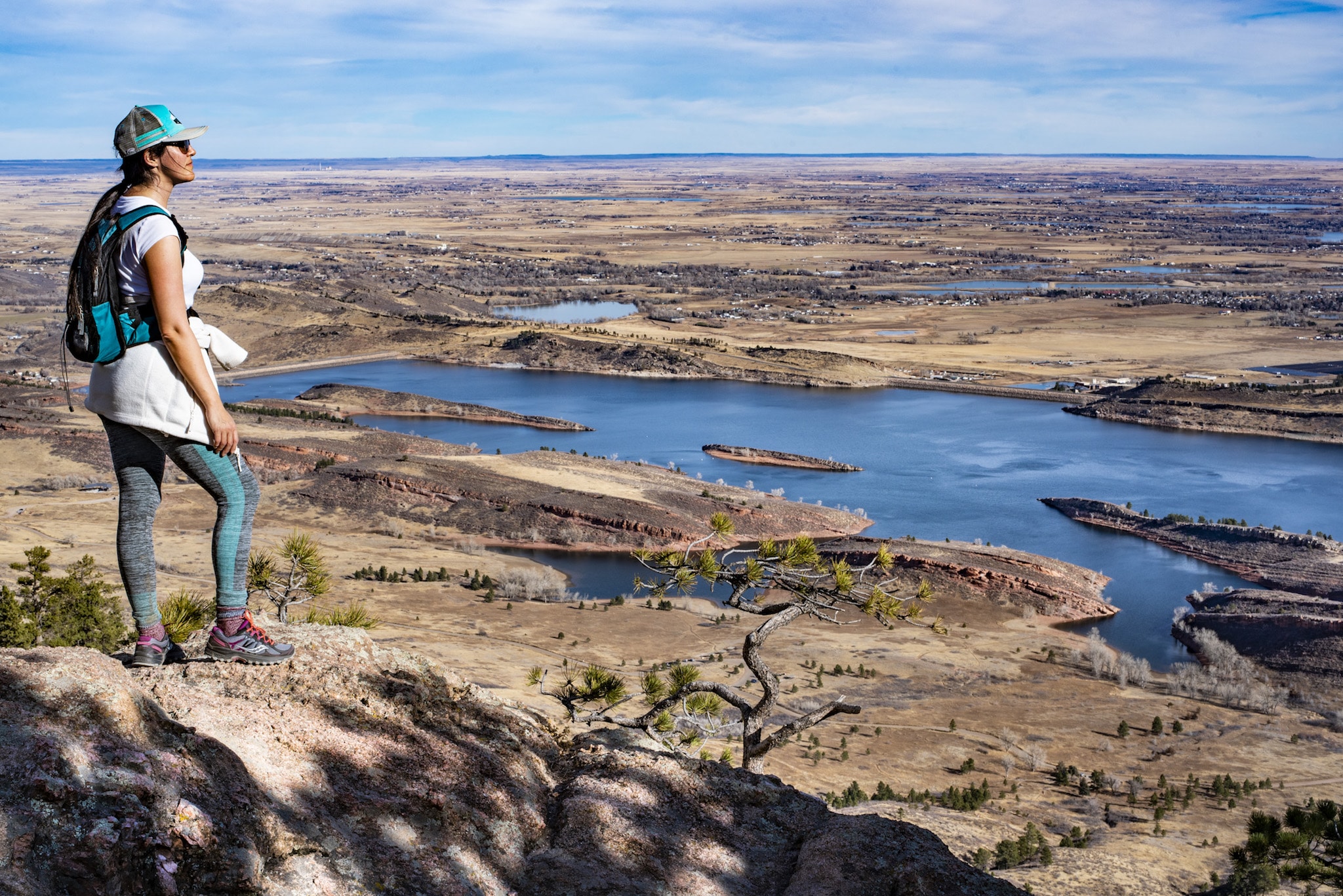 A female hiker gazing at the yellowed landscape from a prominent rocky point, multiple lakes and towns are visible below.