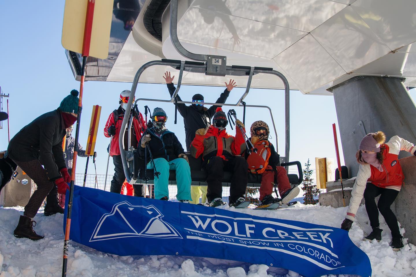First chairlift at opening day at Wolf Creek Ski Area. Three people sit on a chairlift while employees hold blue Wolf Creek Ski Area banner in front of them